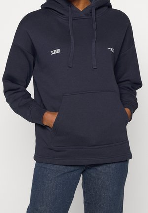 Personne portant un sweat à capuche bleu marine avec des poches avant et un petit texte blanc sur la poitrine, associé à un jean bleu foncé.