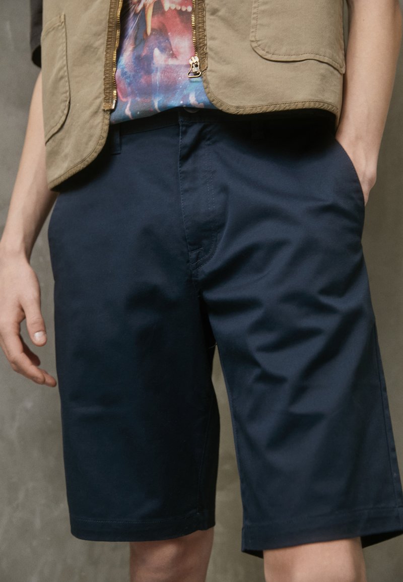 Chinos bleu marine avec une texture lisse, dotés de poches avant et d'une coupe décontractée, associés à un t-shirt graphique à motifs sous un gilet beige.