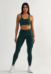 Ciemnozielony stanik sportowy o krzyżującym się designie, zestawiony z pasującymi leginsami z wysokim stanem. Oba elementy mają gładką, elastyczną teksturę.