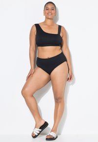 Zwart geribbeld bikini-set met een top met één schouder en een high-waisted onderkant. Model draagt witte sandalen met zwarte zolen. Glad, textuurstof.