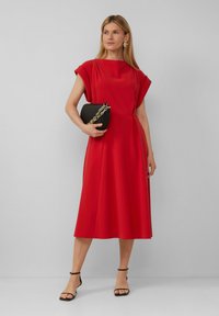 Robe rouge à coupe décontractée et manches courtes, arrivant au milieu du mollet. Associée à une pochette noire avec des accents en chaîne dorée.