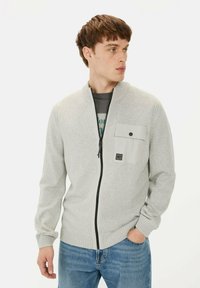 Lichtgrijze zip-up trui met hoge kraag, textuur breiwerk en een borstzak met drukknoopsluiting. Gecombineerd met blauwe spijkerbroek.