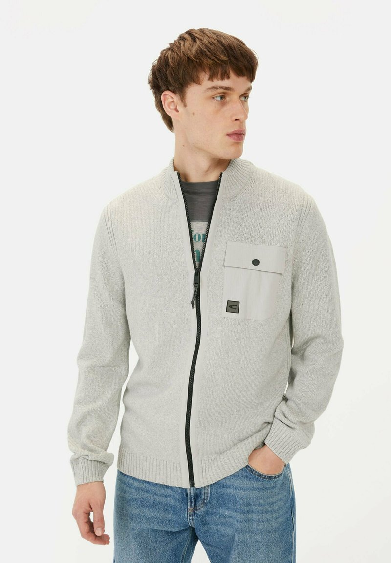 Lichtgrijze zip-up trui met hoge kraag, textuur breiwerk en een borstzak met drukknoopsluiting. Gecombineerd met blauwe spijkerbroek.