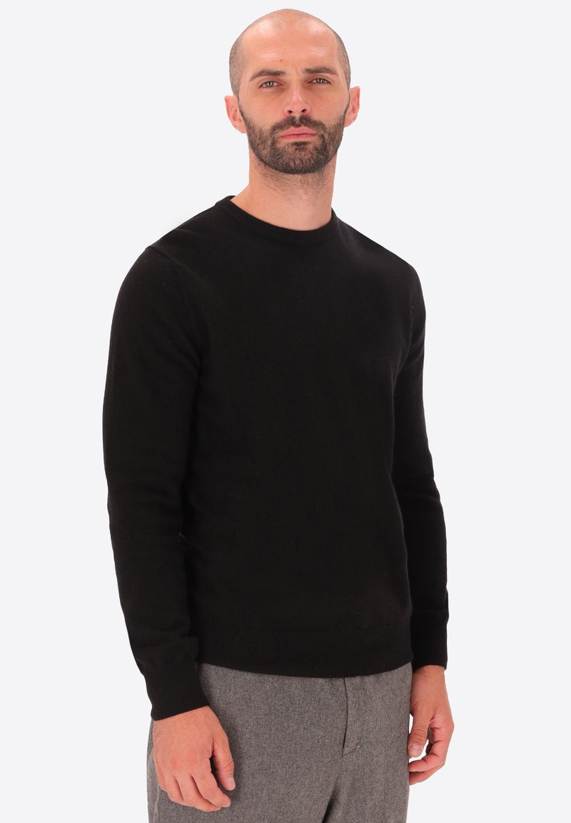 Uomo con barba che indossa un maglione nero a maniche lunghe e pantaloni grigi, in piedi su uno sfondo chiaro e uniforme.