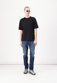 T-shirt noir à manches courtes avec des coutures rouges, jeans en denim bleu et baskets grises. Les accessoires incluent des lunettes de soleil blanches, fines et angulaires.