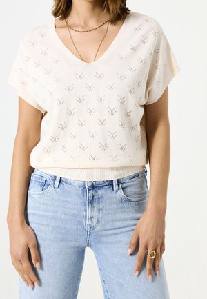 Vrouw die een crèmekleurige korte mouw gebreide top met een V-hals draagt en lichtblauwe high-waisted jeans, geaccentueerd met gouden kettingen en een dikke ring.