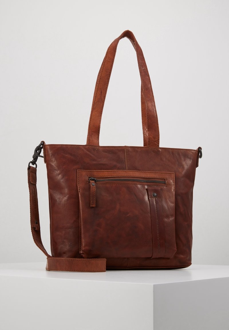 Sac cabas en cuir marron avec doubles poignées, poche avant zippée et bandoulière amovible posé sur une surface blanche.