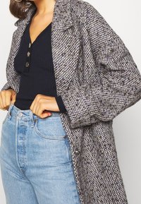 Grijs-witte jas met houndstooth-patroon, voorzien van een kraag, gestructureerde schouders en lange mouwen, gecombineerd met een donkere geribbelde top en licht denim.