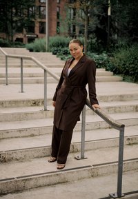 Woman wearing a brown coat and trousers leaning on a metal handrail on concrete outdoor stairs with green plants in the background.
