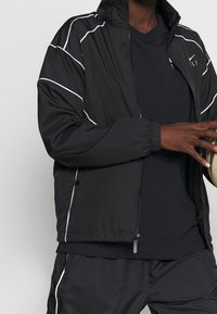 Chaqueta deportiva negra con ribetes blancos, cierre de cremallera y puños elásticos. Lleva una camisa negra debajo y sostiene un balón de baloncesto visible en la mano.