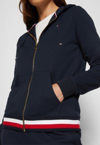 Mujer vestida con una sudadera con cremallera azul marino, con dobladillo a rayas rojas y blancas, cremallera dorada y la mano en el bolsillo delantero, mostrando solo la parte superior del torso.