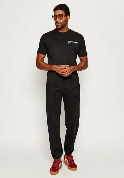 T-shirt nera in cotone con logo ricamato bianco, pantaloni felpa neri e sneakers rosse con strisce bianche. Gli accessori includono occhiali con lenti gialle.