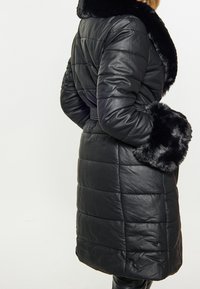 Czarna pikowana kurtka puchowa z kołnierzem i mankietami z sztucznego futra, posiadająca pikowany wzór i pasek w talii. Gładka faktura, długość do kolan.