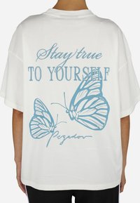 Fehér, túlméretezett póló, kék lepkés grafikával és a "Maradj hű önmagadhoz" felirattal, kézírásos stílusú aláírással.