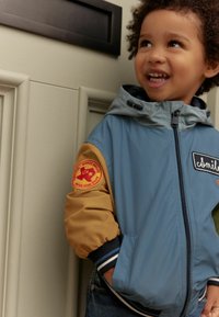Blauwe en beige hooded jas met ribbelmanchetten, een patch op de mouw en een rits aan de voorkant. De stof lijkt glad, met een contrasterend ontwerp.