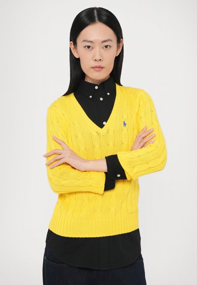 Dzeltens džemperis ar adījuma rakstu un V veida kaklu, valkāts pāri melnai kreklam. Džemperim ir ribota aproce un smalka logotipa detaļa.