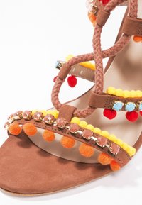 Sandale en daim marron avec des brides tressées, décorée de pompons jaunes, de boules duveteuses orange et d'accents de pierres précieuses colorées.