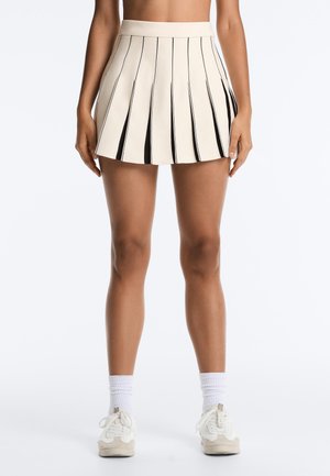 Vrouw die een crèmekleurige geplooide minirok met zwarte binnenpanelen draagt, witte sportsokken en witte sneakers, staand tegen een effen achtergrond.