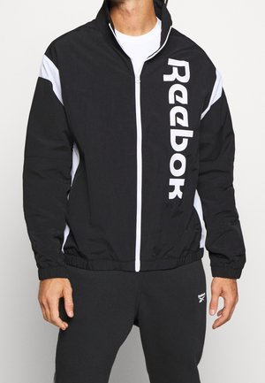 Mann trägt schwarze und weiße Reebok Trainingsjacke mit vertikalem Logo und passende schwarze Jogginghose mit kleinem Logo.