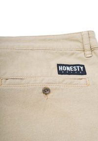 Beiger Baumwollstoff mit einer Paspeltasche; ausgestattet mit einem braunen Knopf und einem schwarzen Etikett mit der Aufschrift "EHRLICHKEIT REGIERT" in weißem Text.