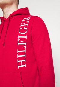 Felpa rossa realizzata in morbido tessuto con tasca frontale, a sinistra presenta la scritta bianca "HILFIGER" e un cappuccio con cordini.