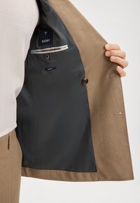 Brauner maßgeschneiderter Blazer mit einer glatten, dunkelgrauen Innenauskleidung. Verfügt über ein Slim-Fit-Etikett und eine kleine Tasche mit gestreiftem Akzent.