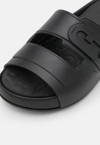 Sandale noire à enfiler avec deux larges brides à découpes, fabriquée en matériau lisse ressemblant à du caoutchouc, avec une marque embossée sur la semelle intérieure.