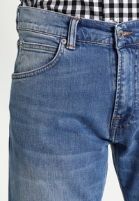 Light blue denim jeans with a faded finish, five-pocket design, and contrast stitching; worn with a black and white checkered shirt.
