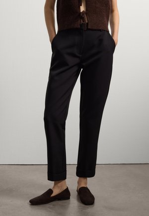 Pantaloni neri su misura con una texture liscia, pieghe ai polsini e tasche laterali. Indossati con un top in maglia marrone e scarpe scivolate scure.