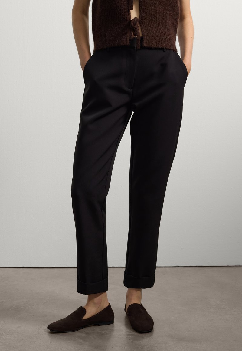 Pantaloni neri su misura con una texture liscia, pieghe ai polsini e tasche laterali. Indossati con un top in maglia marrone e scarpe scivolate scure.