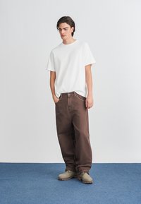 Hvit t-skjorte med korte ermer, brune baggy bukser og beige fottøy. Stoffet ser glatt ut med en avslappet passform, passende for avslappet bruk.
