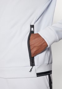 O pulôver de fleece cinza-claro apresenta zíperes pretos, bolsos e um padrão texturizado na lateral. Destaca-se com uma bainha preta em contraste.