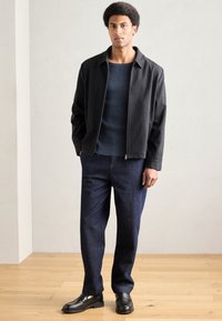 Čierna mikina na zips cez textúrovaný námornícky sveter a tmavomodré nohavice, doplnené čiernymi topánkami, na svetlom drevenom podlahe.