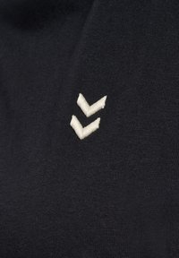 Sort tekstil med en ribbet tekstur, der har et lille hvidt broderet logo med to chevronformer.