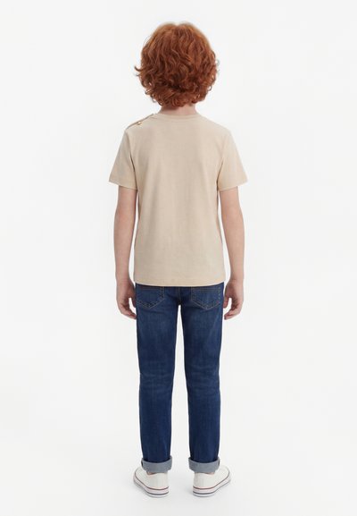 Dieťa s kučeravými červenými vlasmi stojí otočené chrbtom, oblečené v béžovej krátkosrstej košeli, modrých džínsach s vyhrnutými nohavicami a bielych teniskách.