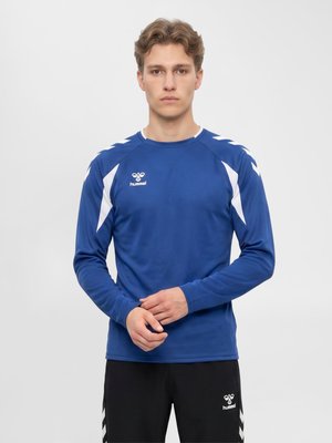Młody mężczyzna w niebieskiej sportowej koszulce z długim rękawem i czarnych spodniach z białymi logo hummel, stojący na jednolitym tle.