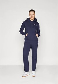 Tmavě modrá mikina s kapucí, klokaní kapsou a stahovací šňůrkou, v kombinaci se sladěnými tepláky. Oba kousky jsou vyrobeny z měkké látky a zdobí je malá značka.