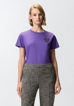 T-shirt di cotone viola con maniche corte e scollo rotondo, arricchita da un design decorativo sul lato sinistro del petto. Indossato con pantaloni con fantasia.
