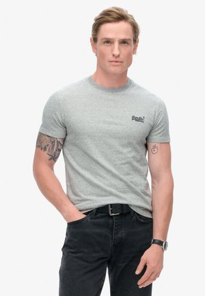 Camiseta gris de manga corta con una textura sutil y un pequeño logo negro en el pecho, combinada con jeans negros y un reloj.