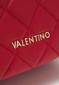 Sac à main en cuir rouge matelassé avec un logo "VALENTINO" doré. La texture est lisse avec des coutures visibles et une forme structurée.