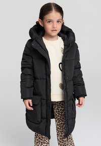 Czarna kurtka puchowa z kapturem, z dużymi przednimi kieszeniami i zapięciem na zamek. Nosi się ją na kremową koszulę z dekoracyjnymi akcentami oraz spodnie w panterkę.