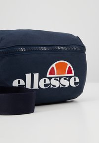 Borsa in tessuto blu navy con chiusura a zip, caratterizzata da un logo bianco e un design di accento arancione. Include una tracolla testurizzata per il trasporto.