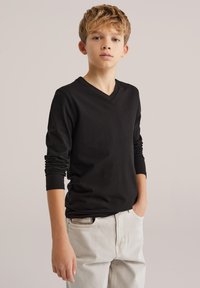 Fekete, hosszú ujjú V-nyakú póló, puha pamut anyagból, világosszürke hétköznapi nadrággal párosítva. A modell zsebre tett kézzel áll.