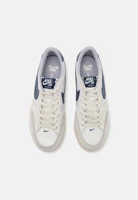Hvite sneakers med marineblå detaljer, med overdel i semsket skinn og mesh, rund tå og flate lisser. Nike-logo på siden og tungen.