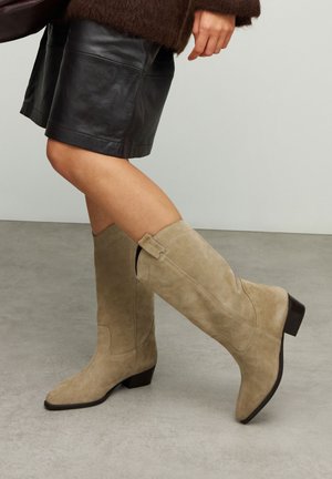 Personne portant des bottes en daim beige montant jusqu'aux genoux et un short en cuir noir, debout sur un sol gris contre un mur uni.