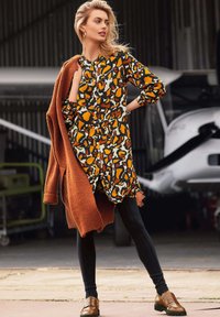 Gestreepte jurk in zwart, oranje en roomkleur, gecombineerd met een gestructureerde rustkleurige cardigan. Zwarte leggings en bruine loafers maken de outfit compleet.