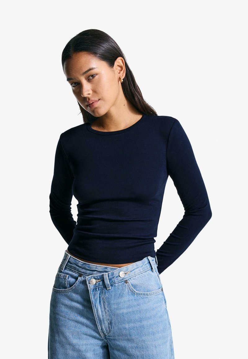 Navy long-sleeve top with a fitted silhouette and a round neckline, made from smooth fabric, paired with light blue high-waisted jeans.