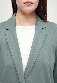 Lysegrønn blazer med spiss krage, myk tekstur og løs passform, brukt over en hvit ribbet topp. Fokuser på overkroppsvinkelen.