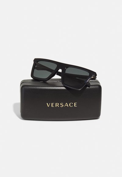 Occhiali da sole neri rettangolari con lenti grigie su un astuccio nero. L'astuccio presenta la scritta "VERSACE" in rilievo con lettering dorato.
