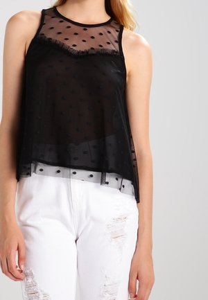 Femme portant un haut sans manches noir transparent à pois superposé à un caraco noir, associé à un jean blanc déchiré.